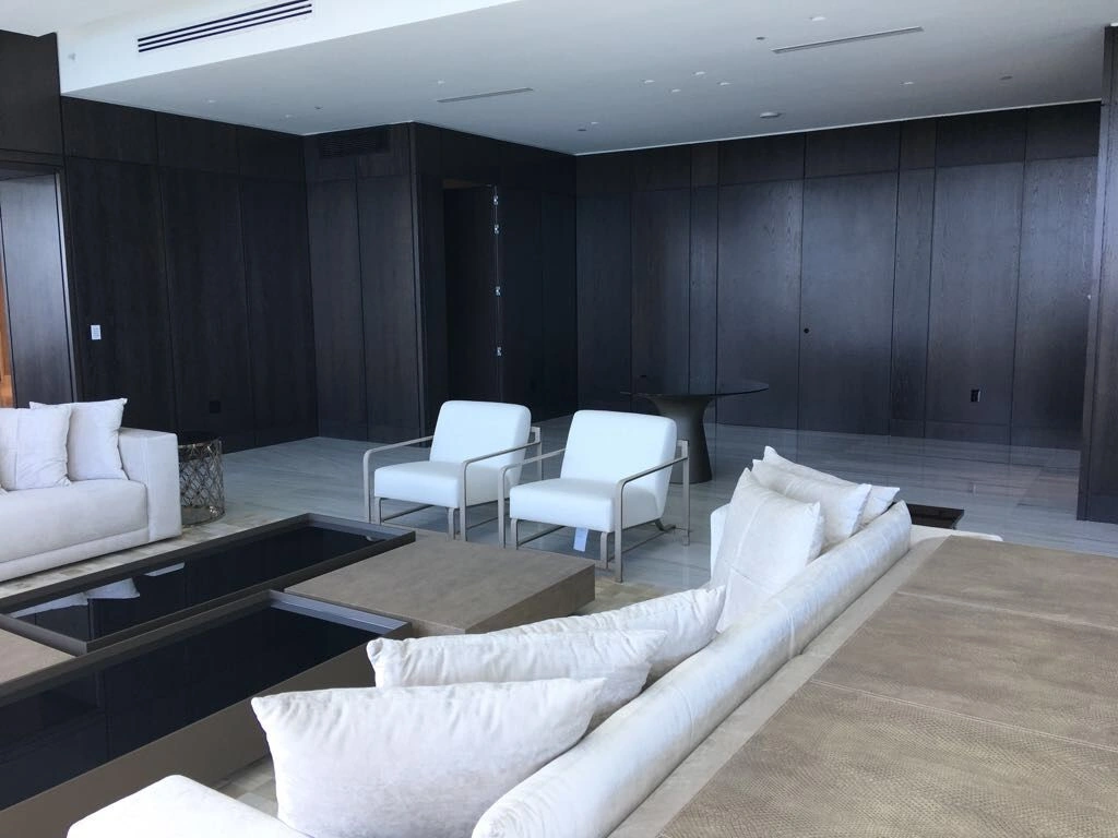Dark wood-grain paneled walls in a lounge with two white club chairs, cream sofas, and low dark glossy tables on light stone flooring