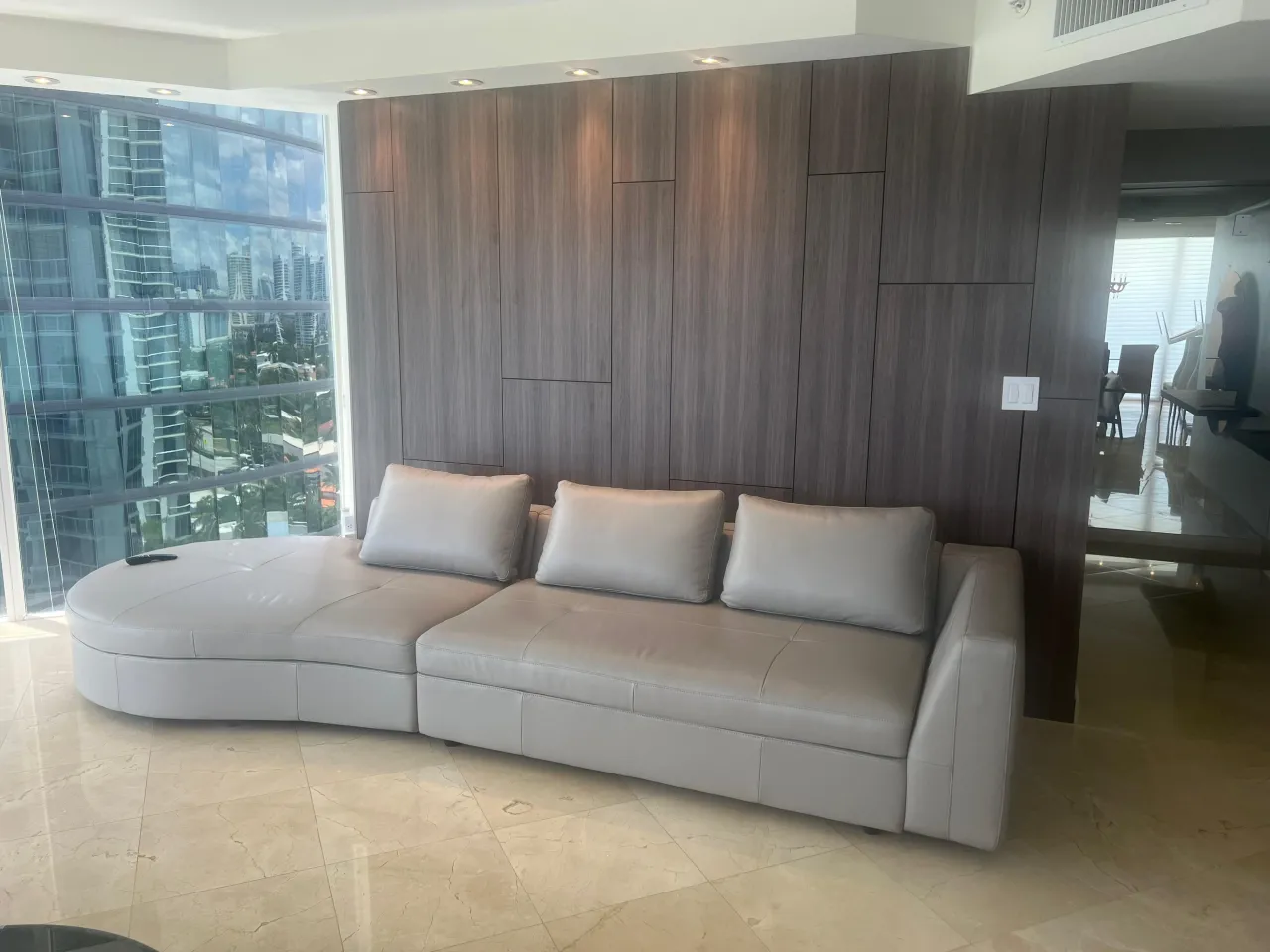 Dark wood-grain panelled accent wall behind a long light gray upholstered sofa, floor-to-ceiling windows with a city skyline view, stone floor