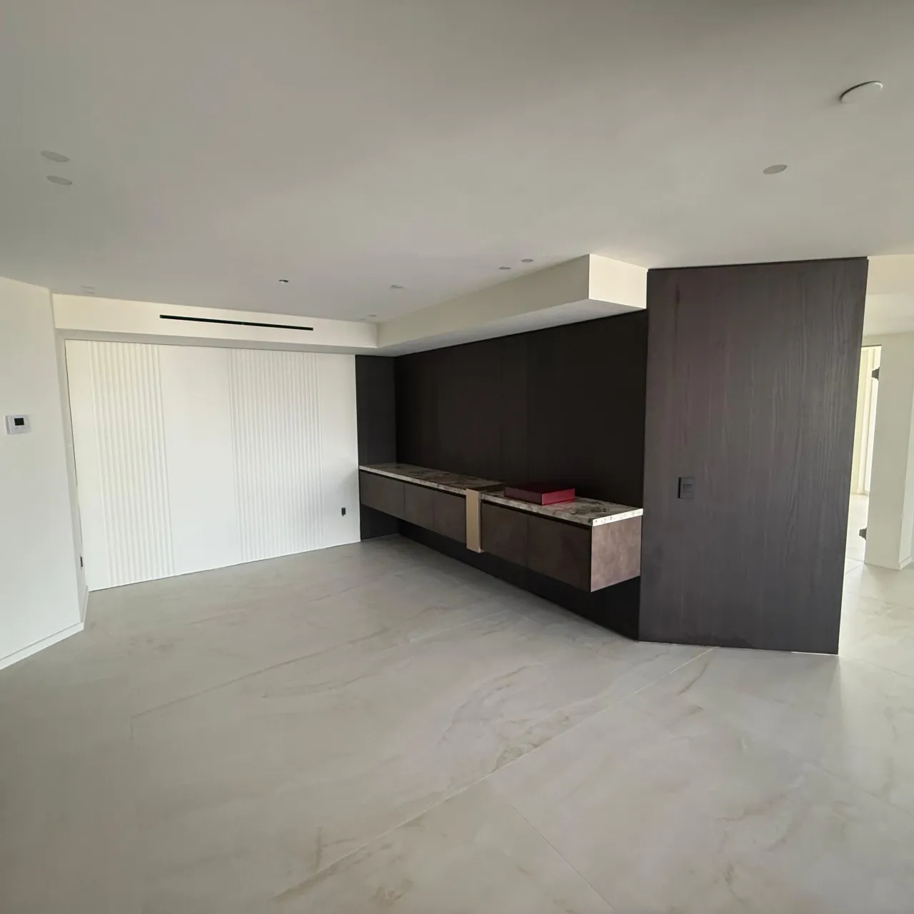 Empty room with a dark wood-grain wall, a floating console run with light stone-look tops, and a wide white fluted panel wall at left, tiled floor