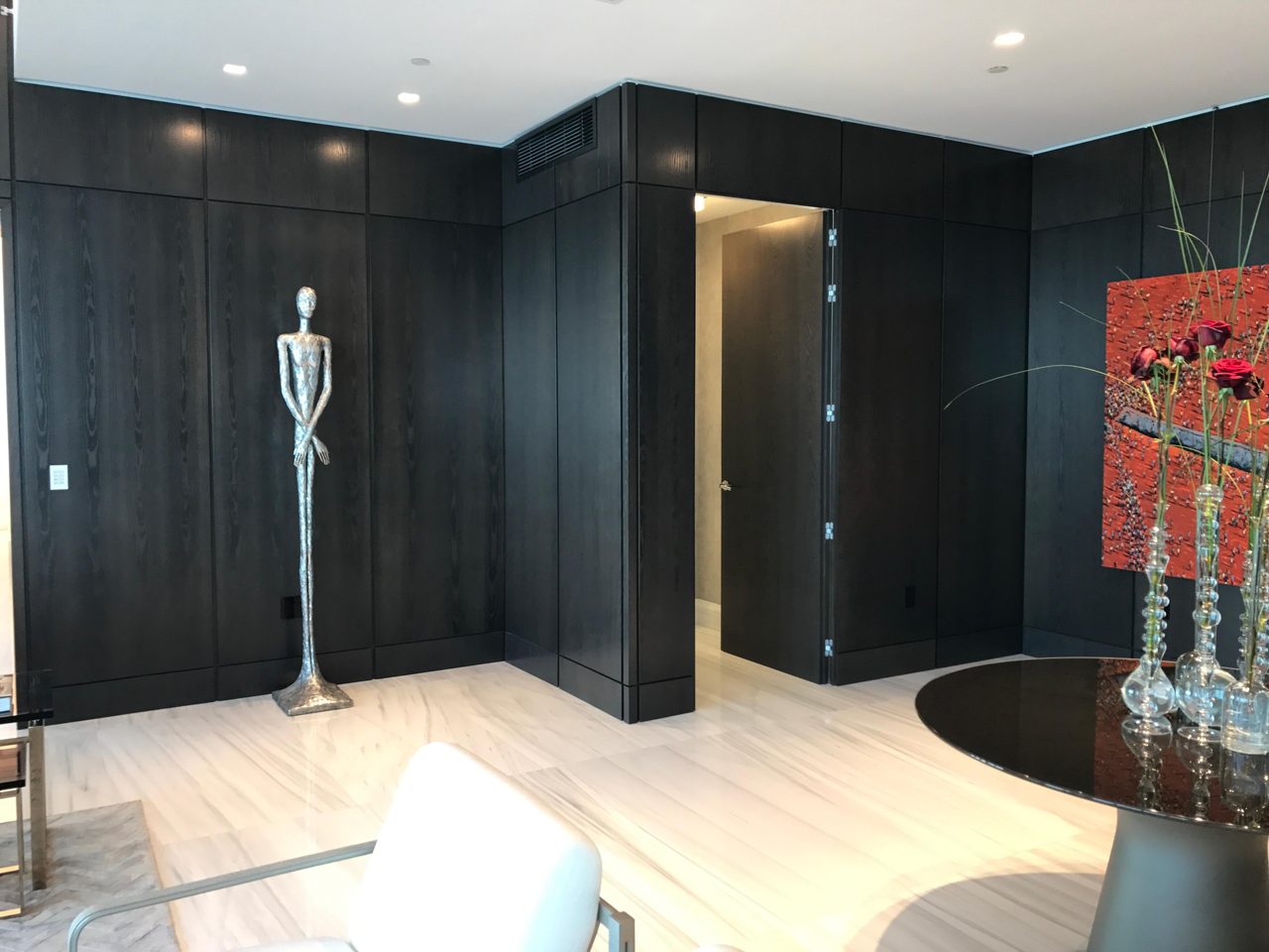 Dark wood-paneled room corner with a standing metal figure sculpture, open door to hallway, round black table with flowers at right