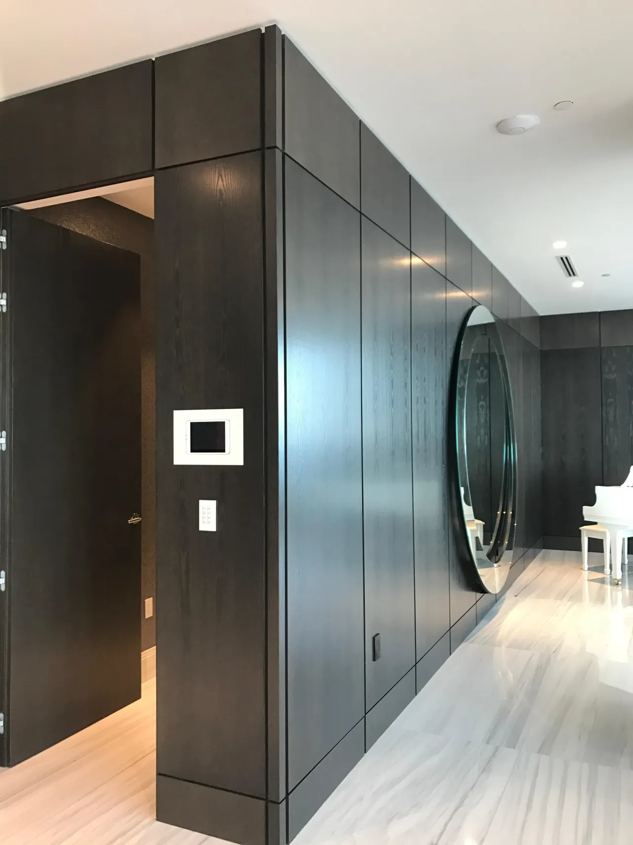 Dark wood-paneled entry wall with an open door revealing a corridor, wall-mounted keypad, large round mirror reflecting a piano, light veined stone floor