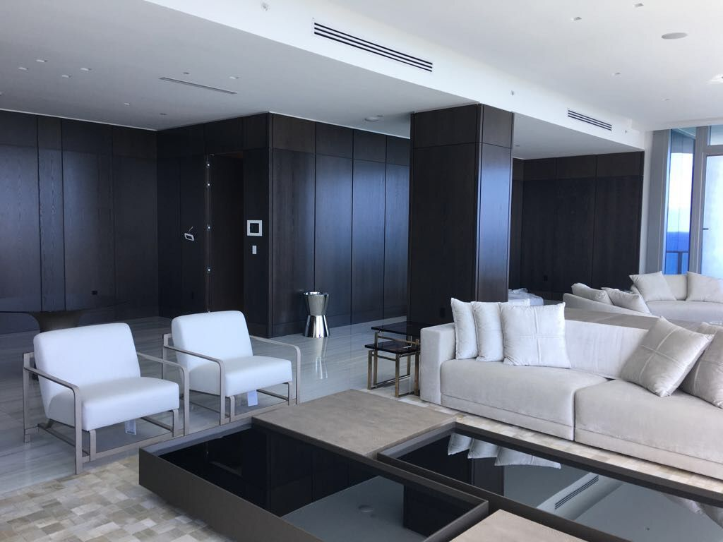 Dark wood-paneled living room corner with white club chairs, black glass coffee tables, and a floor-to-ceiling window view