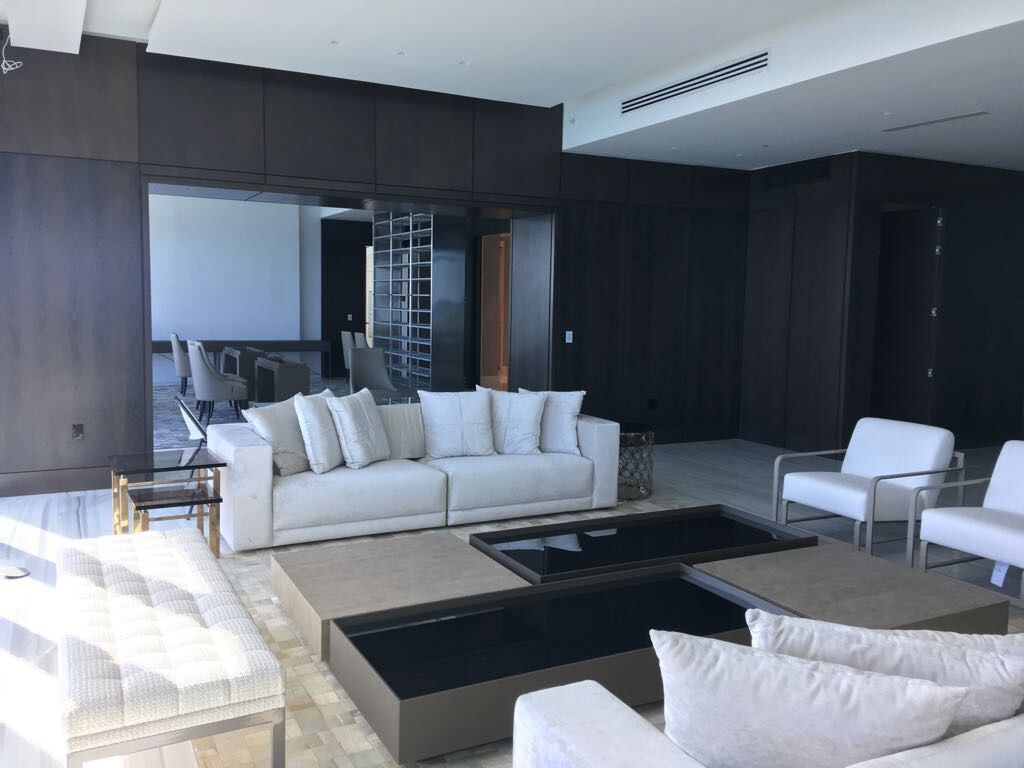 Dark wood-paneled living room with cream sectional sofas, black glass coffee tables, glimpse of dining area through an opening in the paneling