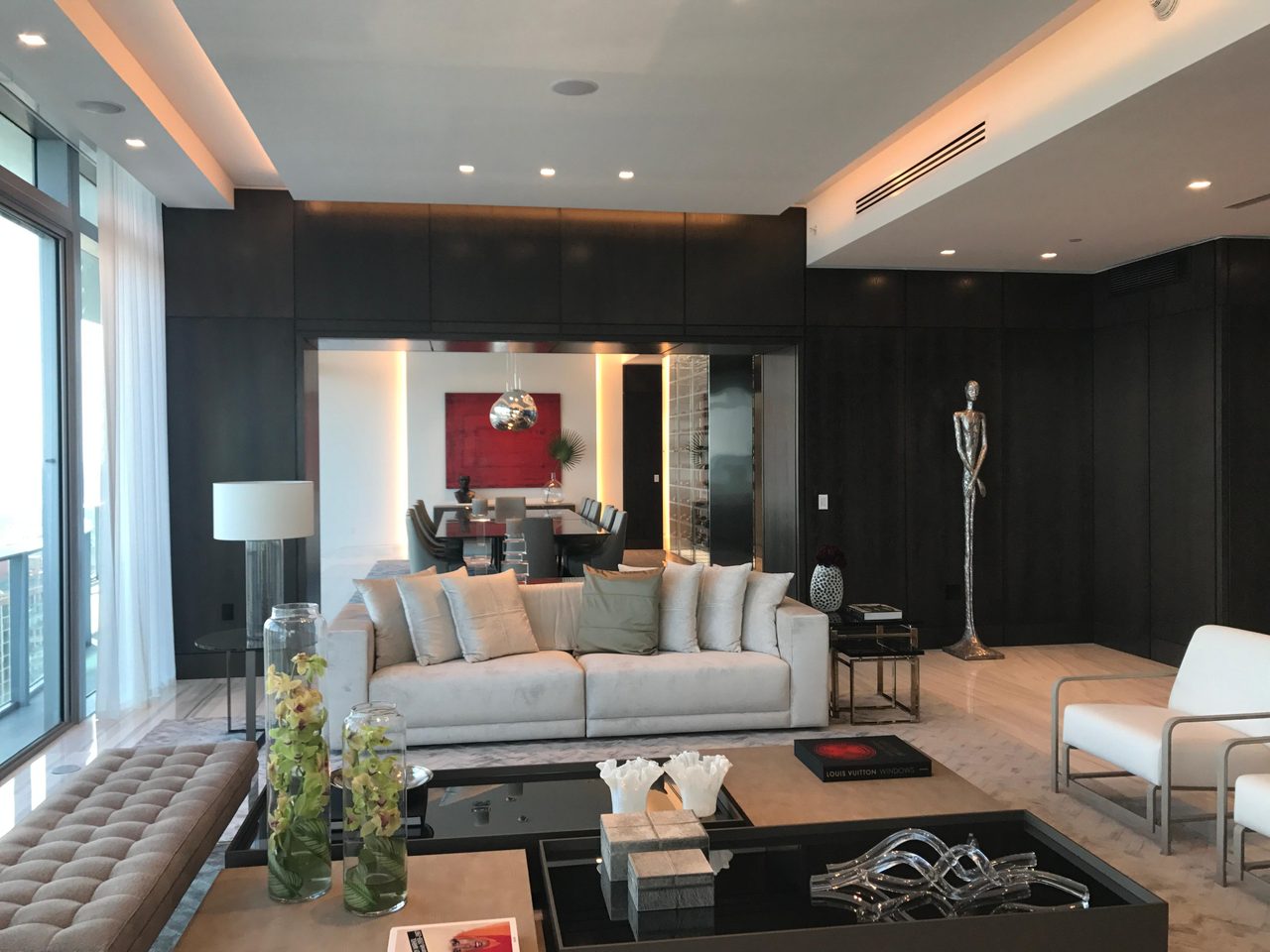 Dark wood-paneled living room with cream sofa, red abstract art accent, standing metal sculpture, floor-to-ceiling city-view windows