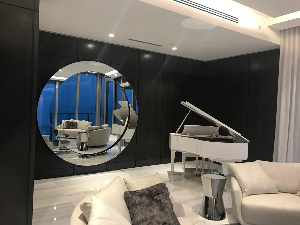 Dark wood-paneled living room wall with a large round mirror reflecting balcony windows, white grand piano beside it