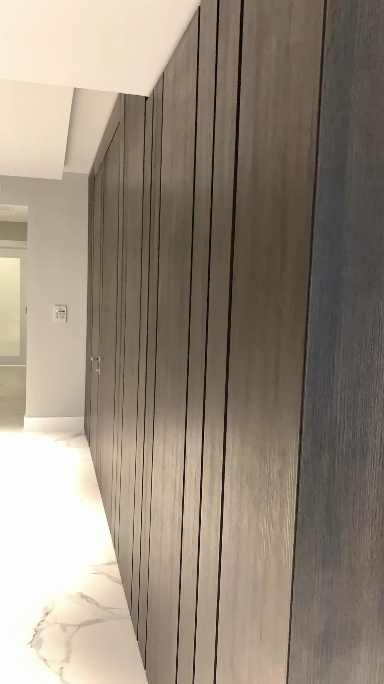 Dark wood vertical slat/board-and-batten wall paneling along a hallway, white veined stone flooring, viewed at an angle