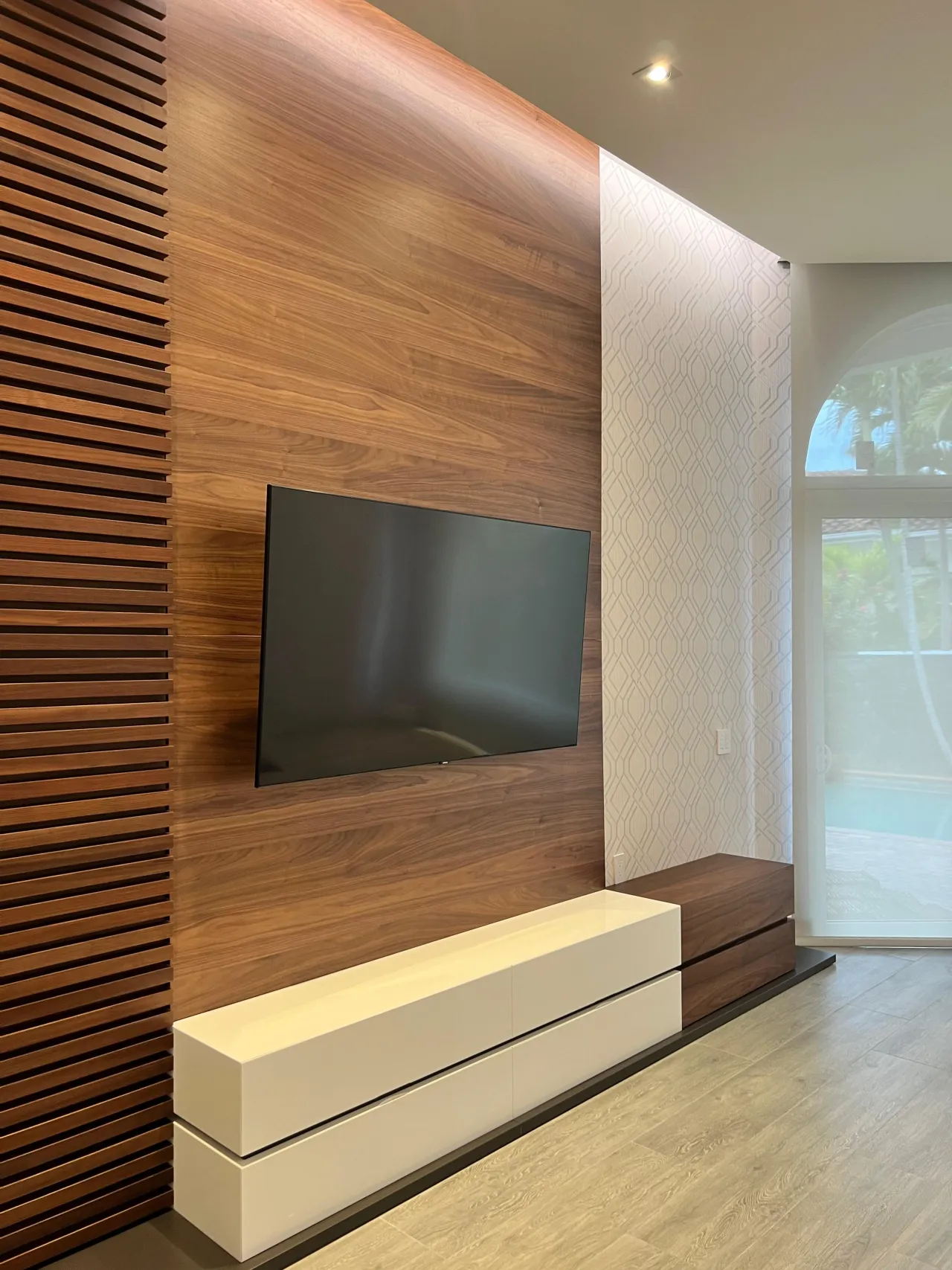Dark wood wall combining vertical slats and a solid panel section, wall-mounted TV, floating console below, geometric wallpaper accent to the side