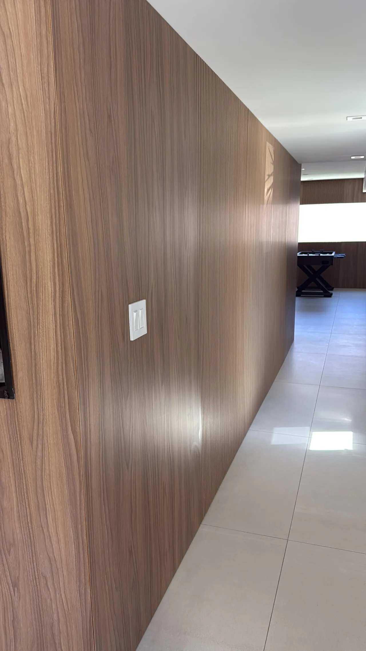 Dark wood wall with vertical fluted detail section and a wall switch plate, transitioning to a tiled hallway floor beyond