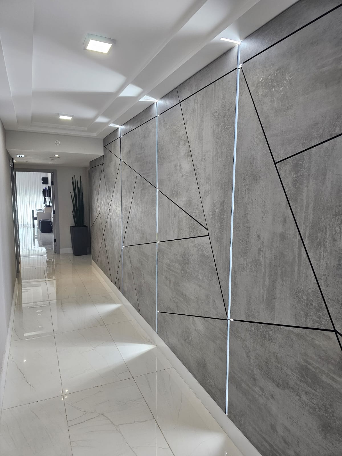 Gray concrete-look panels with black inlay lines and three lit vertical strips, glossy veined tile floor, potted plant at the end of the corridor