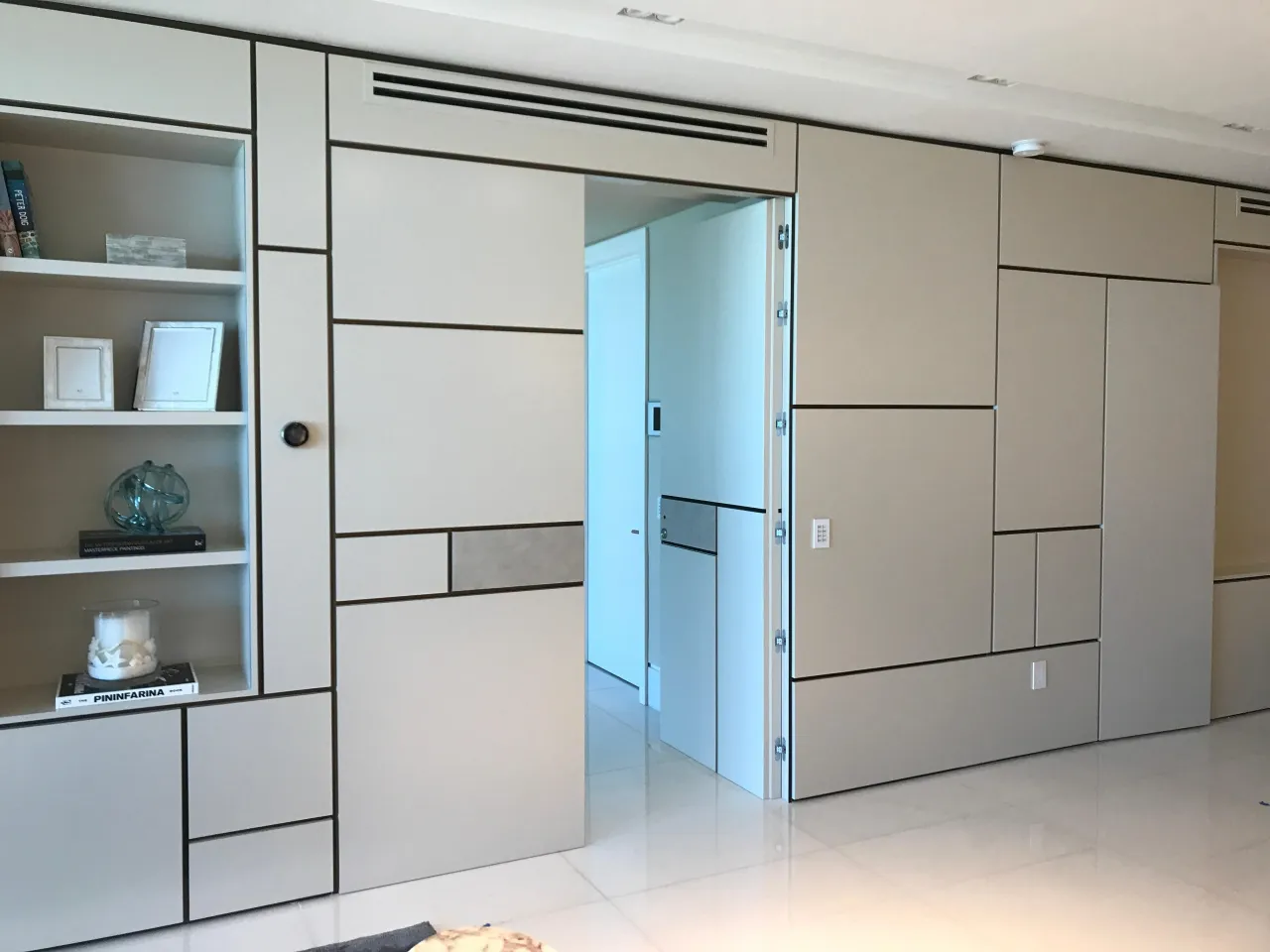 Light cream panelled wall with recessed reveal lines and a built-in open shelf unit holding books and objects, beside an open sliding panel door