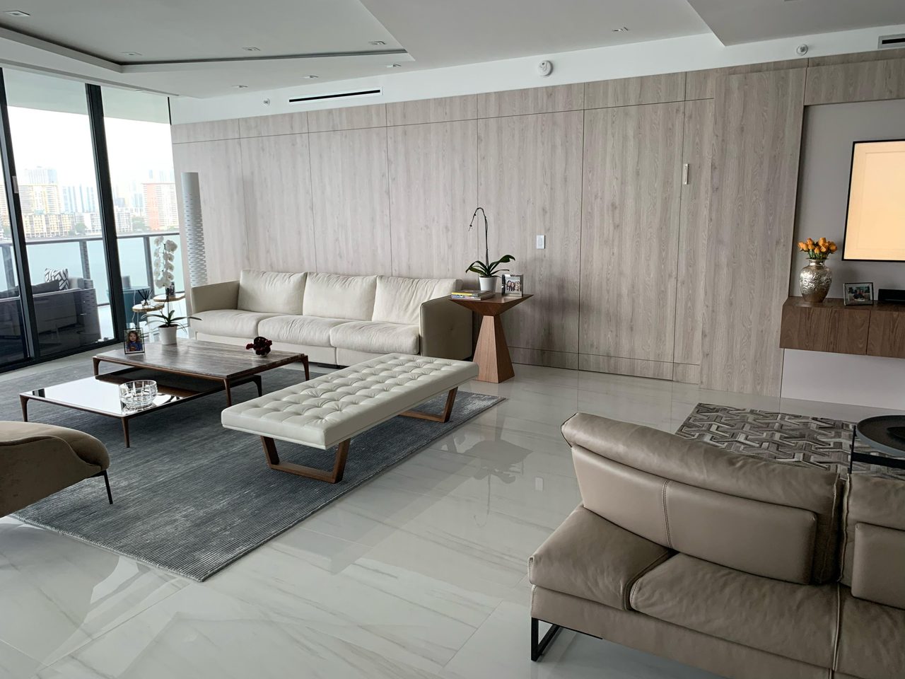 Light gray wood-grain paneled living room wall with flush closet doors, cream sofa, wood side table, city-view windows at left