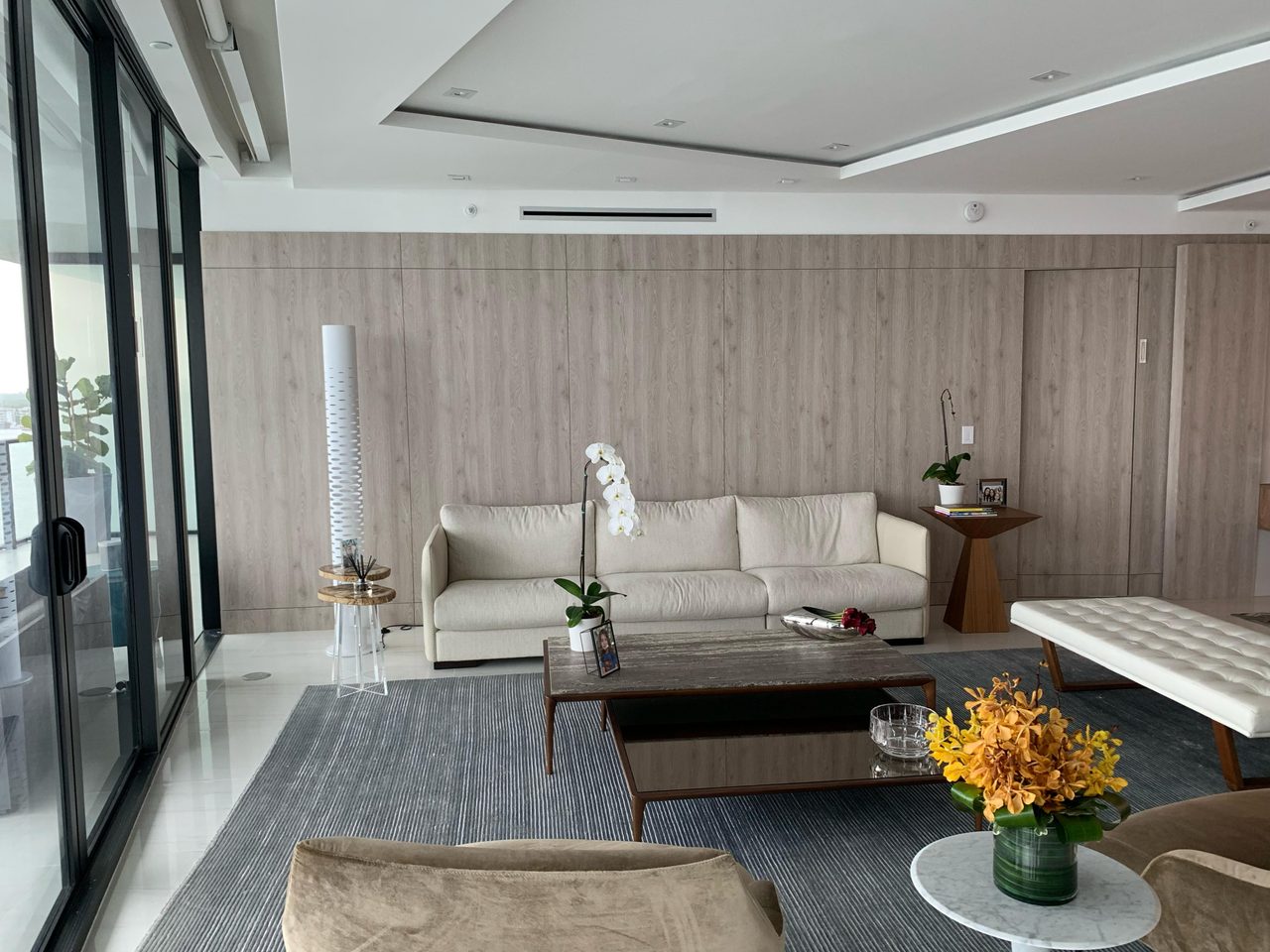 Light gray wood-grain paneled wall behind a cream sofa, with a tall white cylindrical floor lamp and glass balcony doors at left, gray rug in front