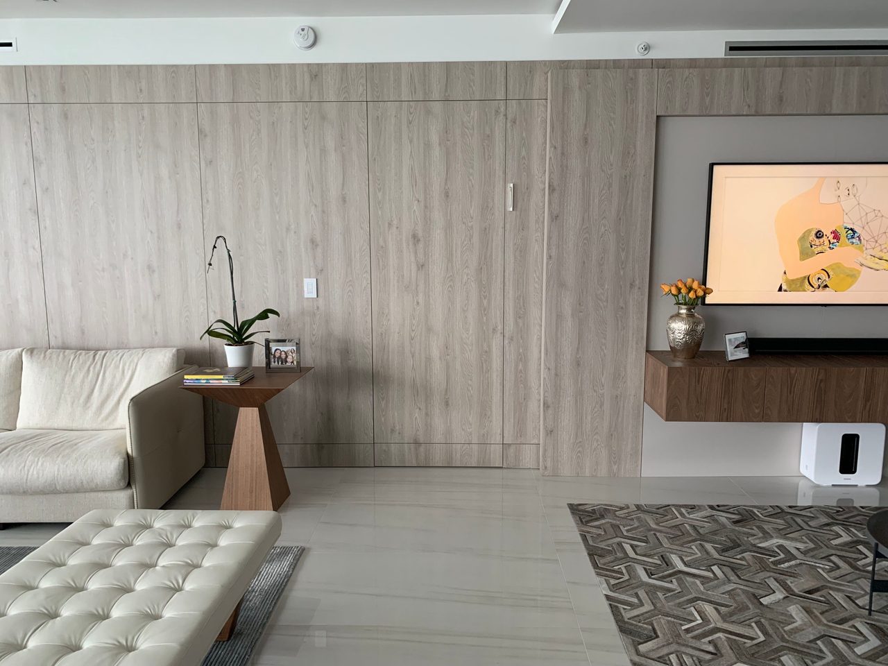 Light gray wood-grain paneled wall with flush closet doors and a wall-mounted TV over a floating wood console, cream sofa and tufted bench