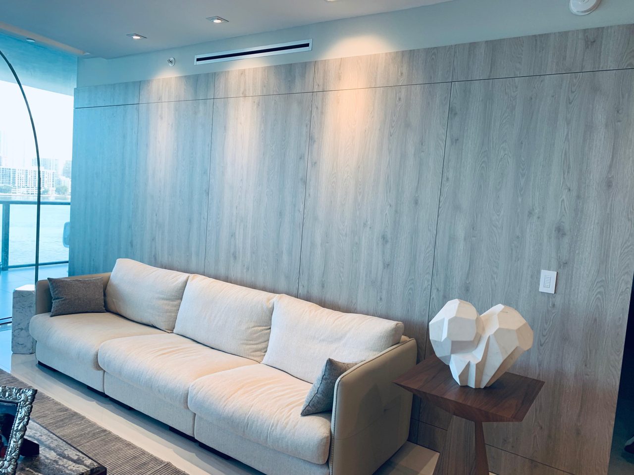 Light gray wood-grain paneled wall behind a beige sofa with sculptural white decor pieces on a small wood side table, waterfront view at left