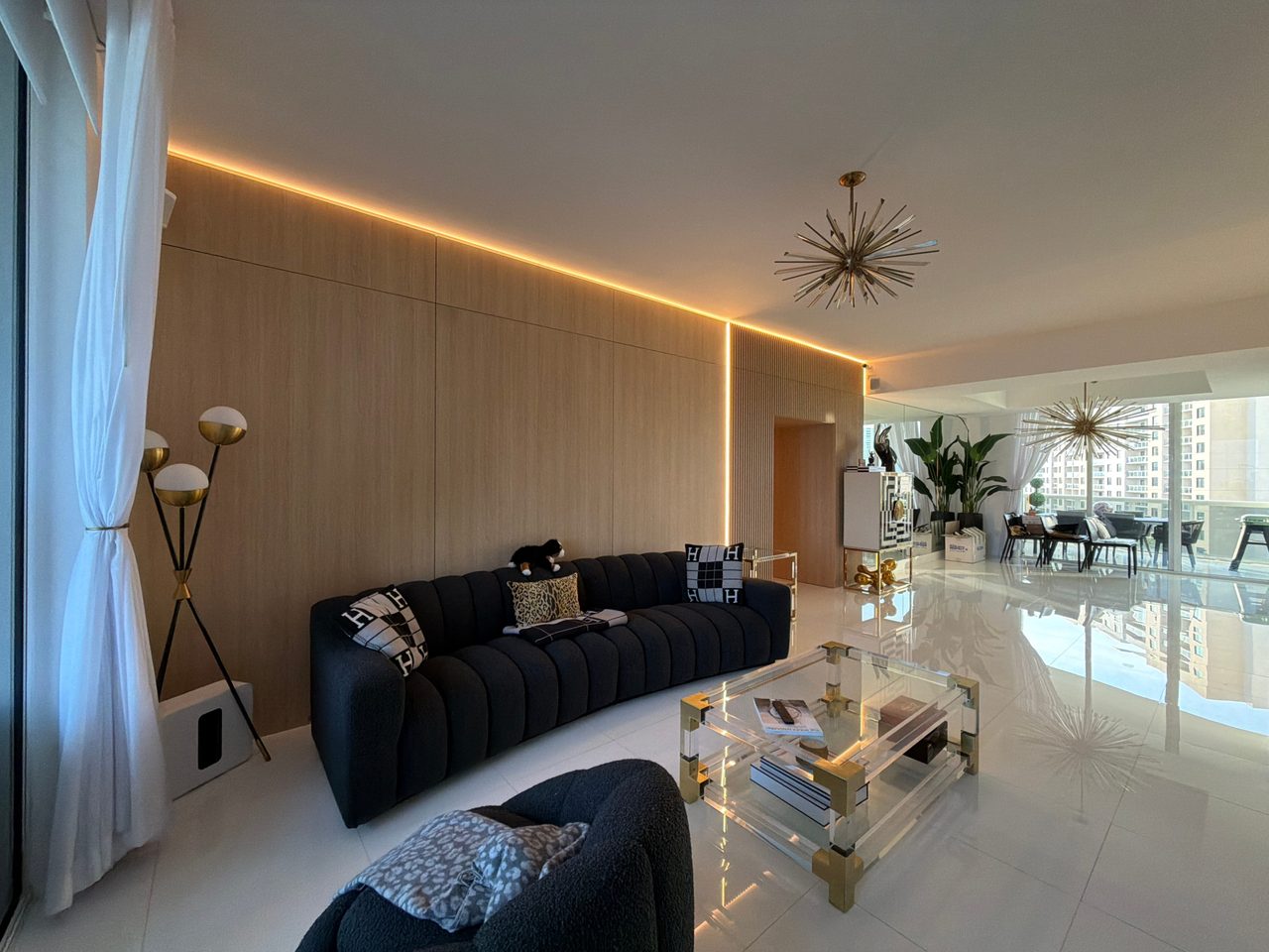 Long light wood-grain flat-panel living room wall with continuous cove lighting above a charcoal sectional, beside floor-to-ceiling windows
