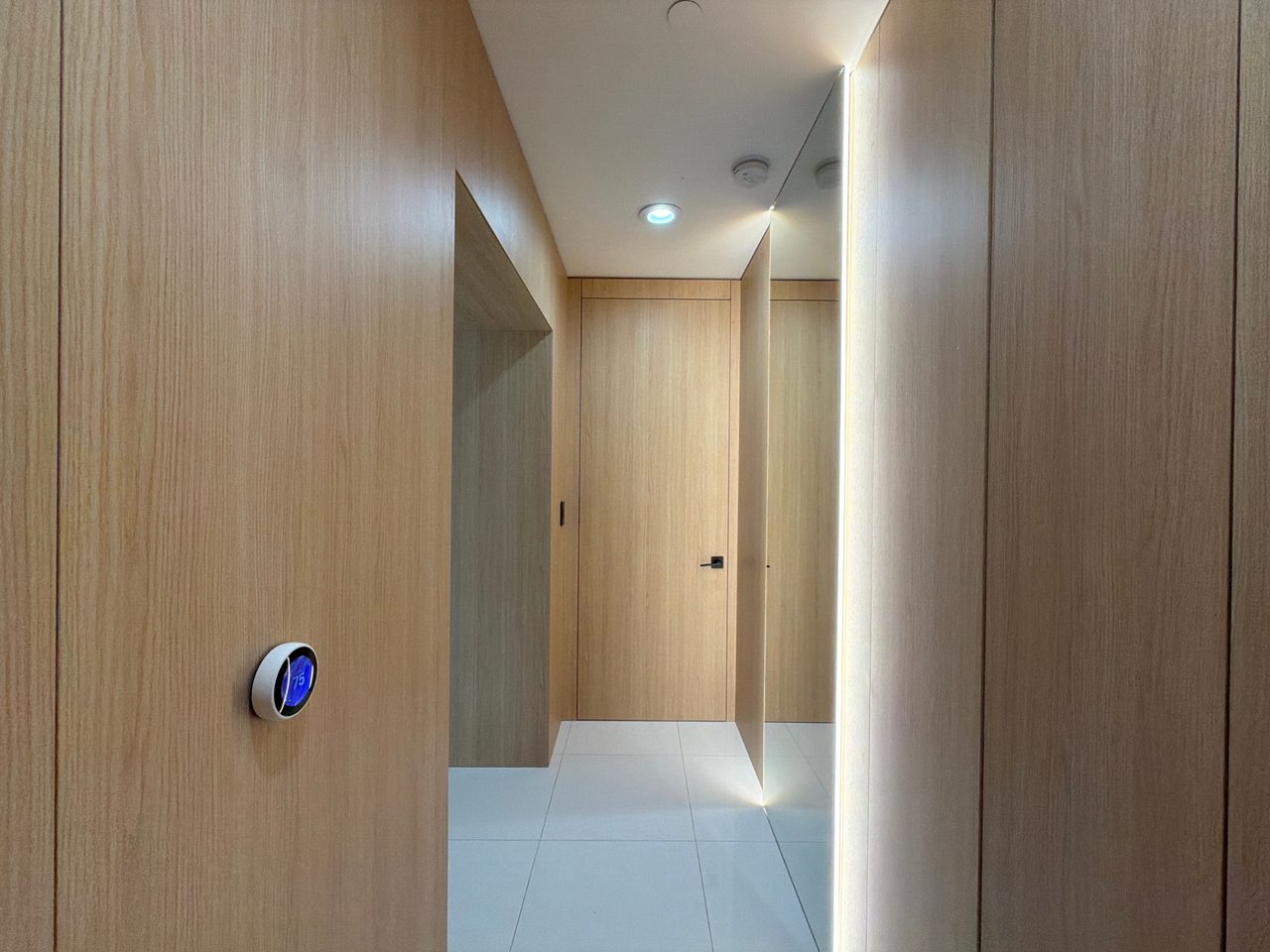 Light wood-grain wall panels with a flush door, a wall-mounted thermostat, and a lit vertical reveal along a hallway on white tile flooring