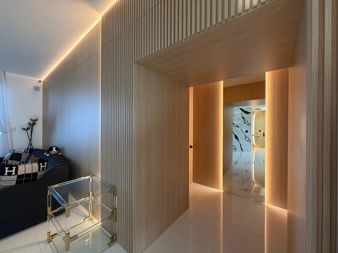 Light wood-grain paneled hallway entry with a slatted wall section above, a lit reveal along the edge, and a flush door at the end of the passage