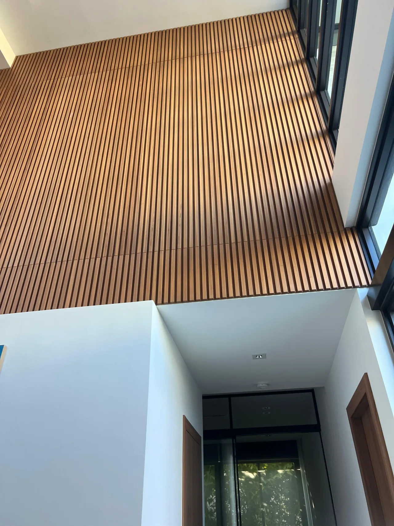 Natural wood slat ceiling and upper wall feature viewed from below in a double-height foyer, with glass door and wood-trimmed doorway visible below