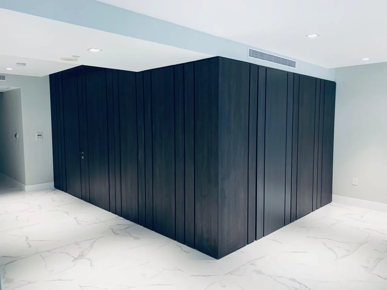 Near-black vertical plank cladding wrapping a freestanding interior volume with a concealed door, set on white veined tile flooring