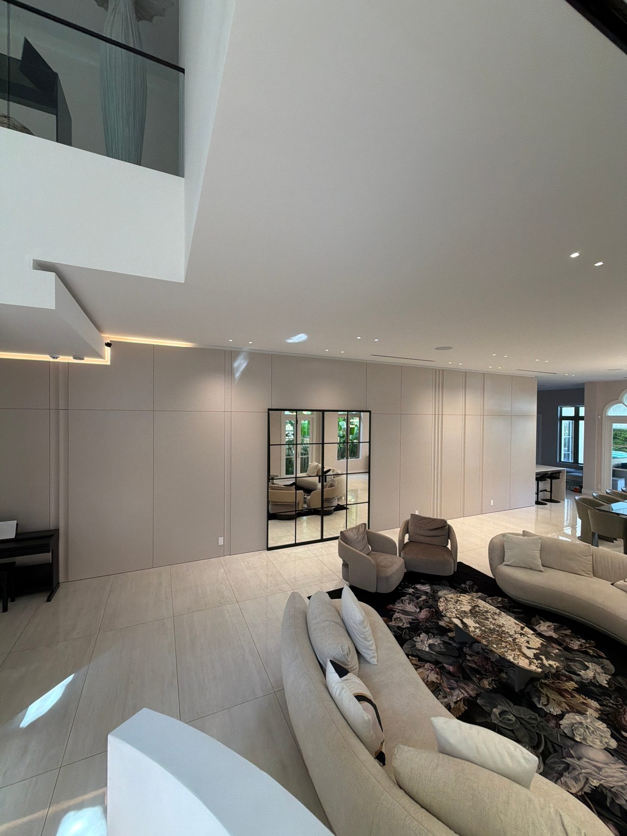 Open room with a long taupe flat-panel wall and black grid-framed mirror, cream curved sofas and floral rug, upper-level glass balustrade above