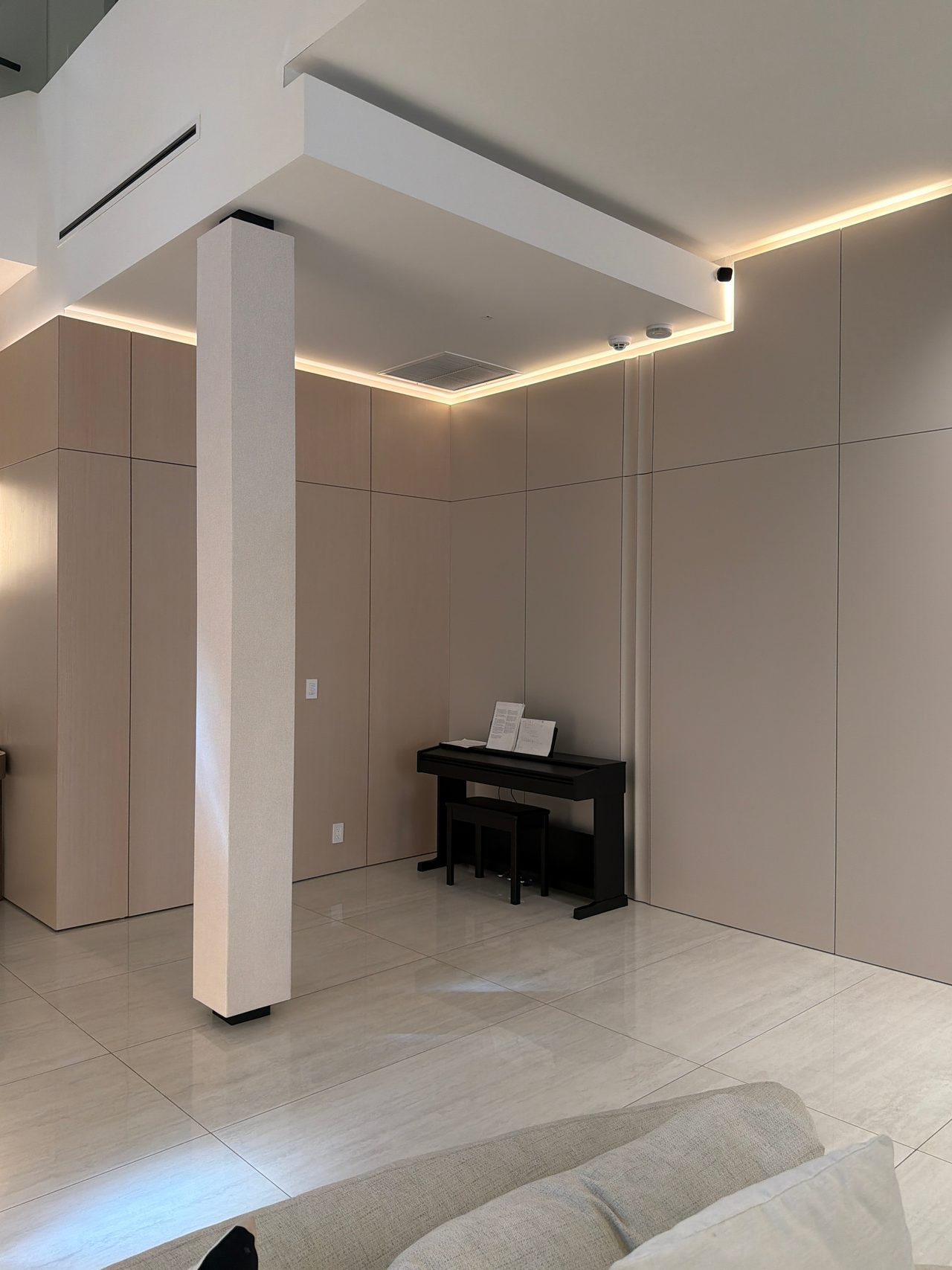 Taupe panel walls meeting at a corner with a white column, cove lighting above, black upright piano and bench, glossy light tile floor