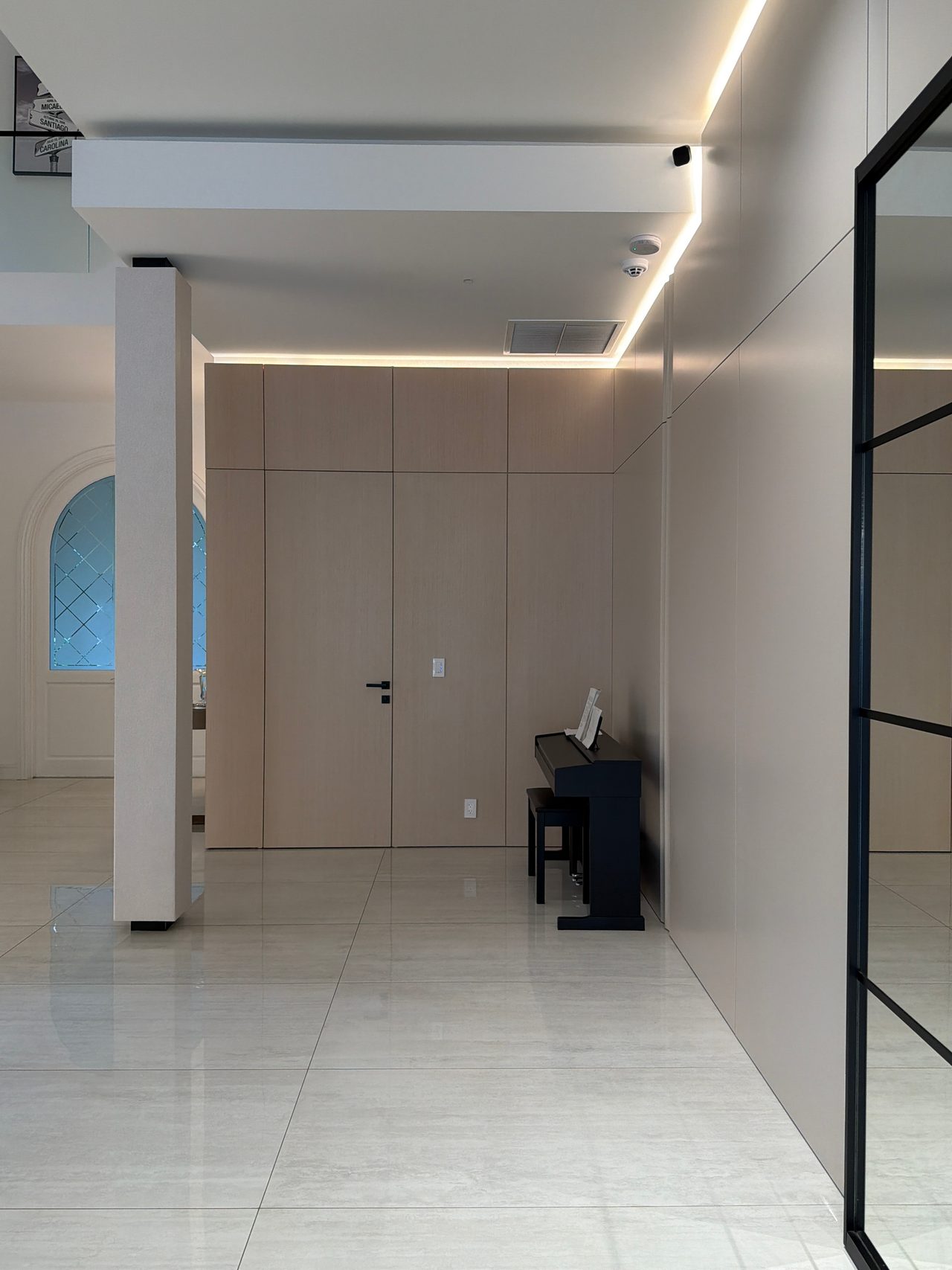 Taupe panel wall with cove lighting and a flush door, black upright piano at the corner, glossy light tile floor, black-framed glass partition at right