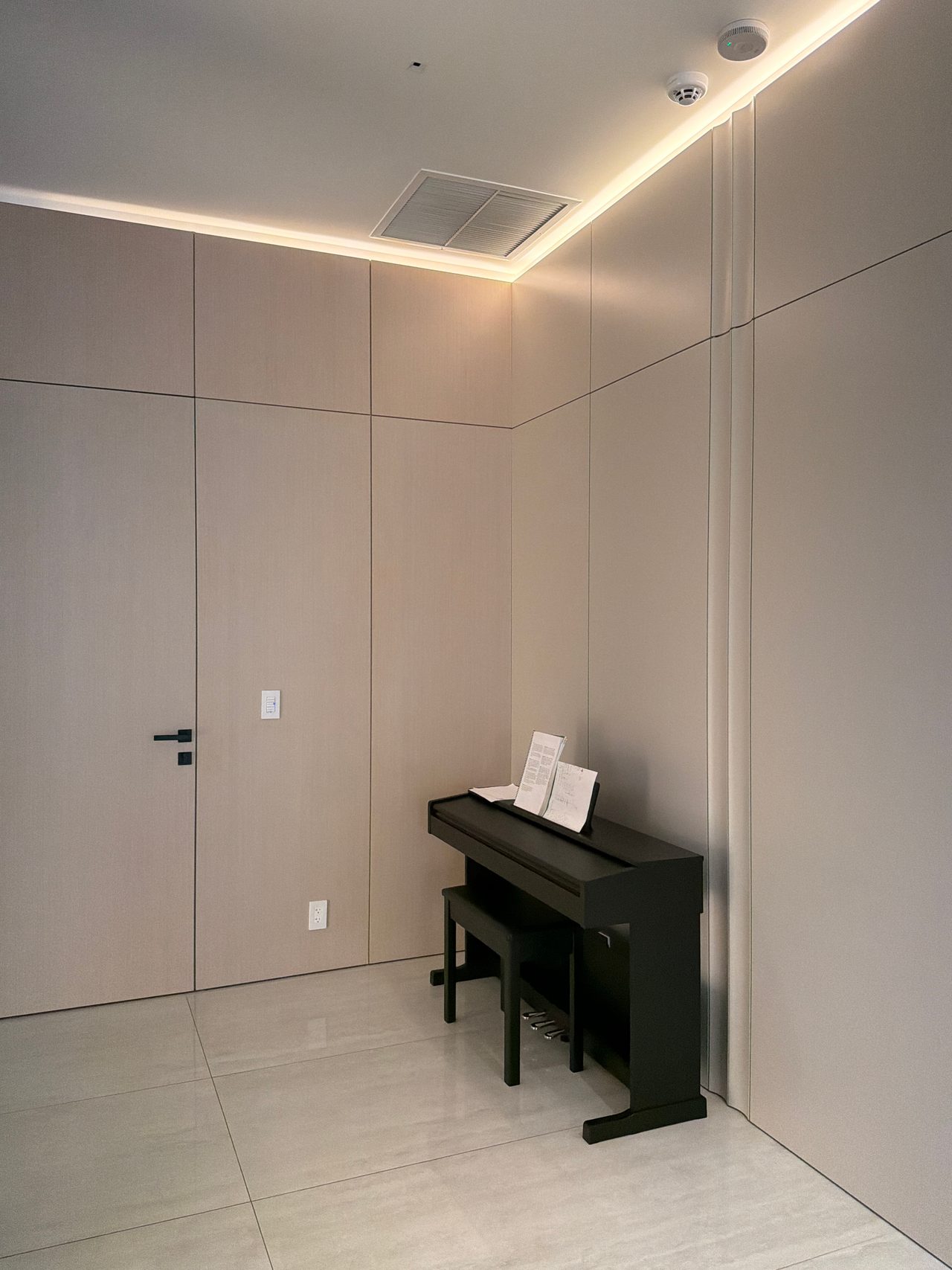 Taupe flat-panel wall alcove with concealed cove lighting, a flush panel door, and a black upright piano with bench on light tile flooring