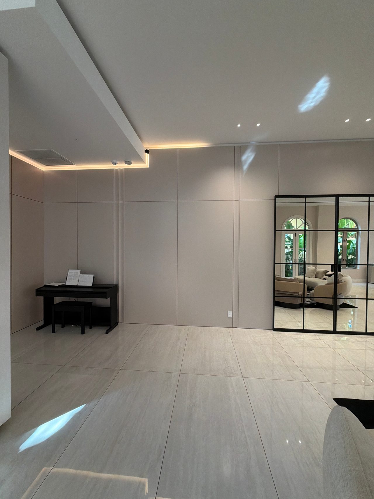 Taupe flat panel wall with cove lighting, a black upright piano in a recess, and a tall black grid-framed mirror reflecting arched windows