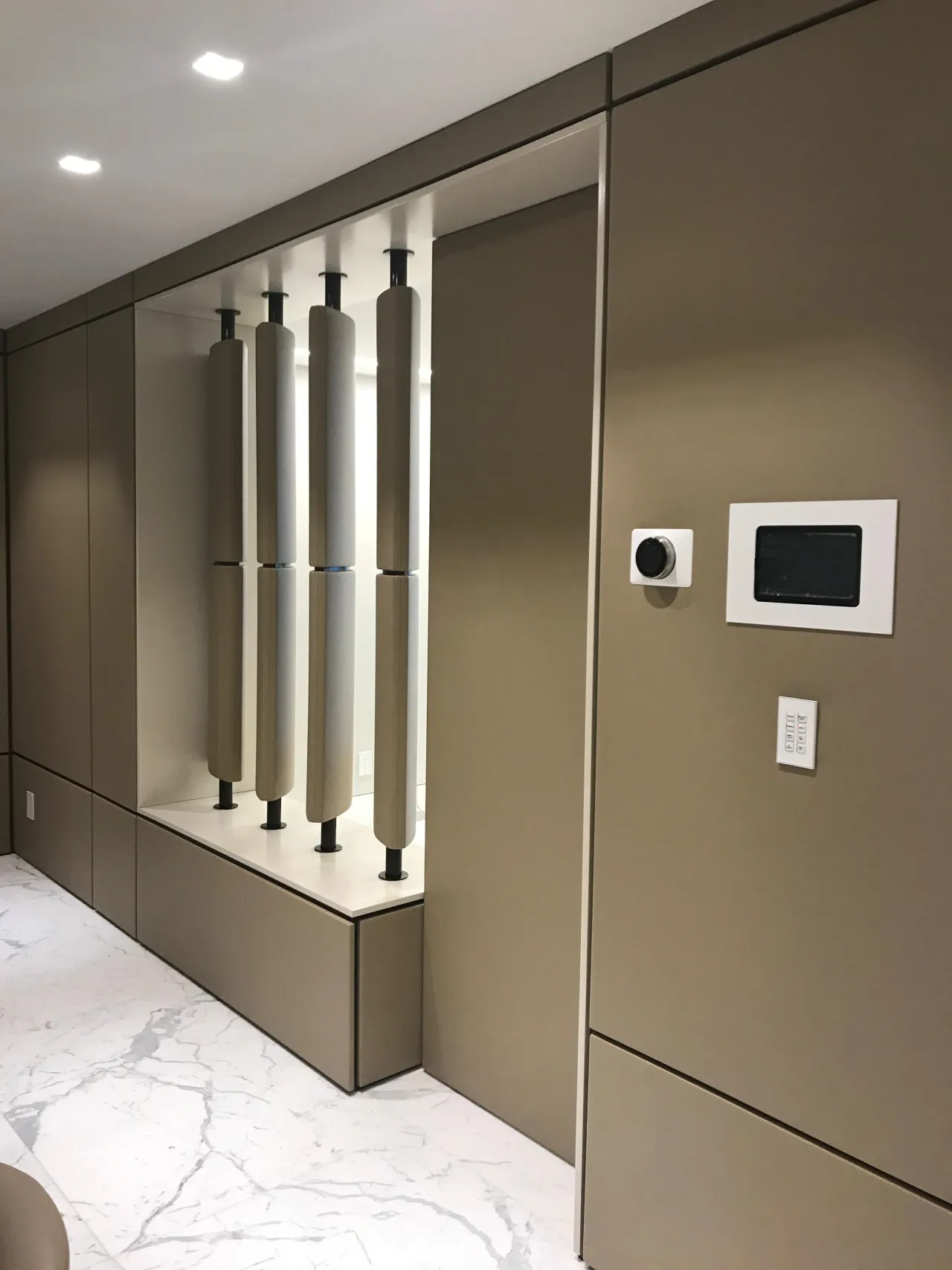 Taupe/khaki cabinet wall with a backlit niche displaying decorative cylindrical objects on a white shelf, wall-mounted tablet controls to the side