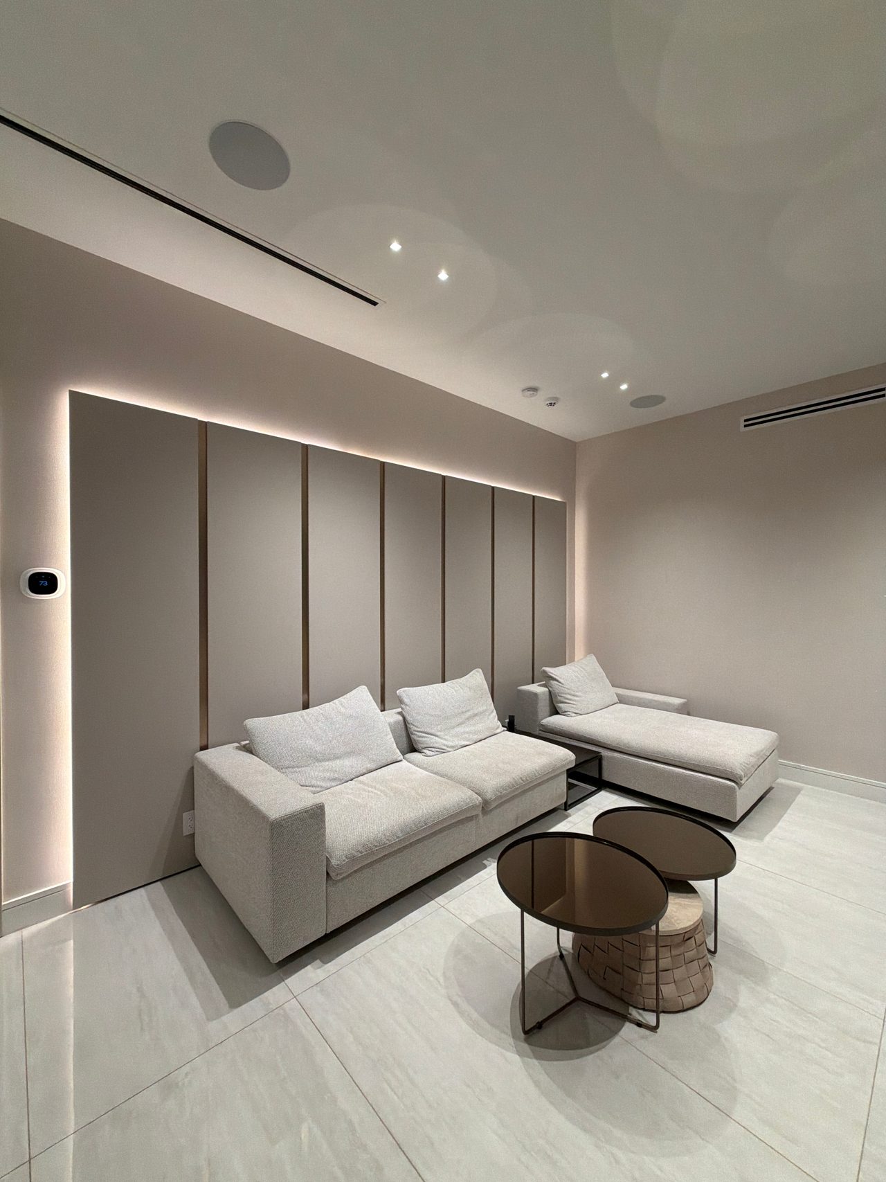 Taupe wall panel with vertical metallic reveal strips and backlit perimeter glow, light upholstered sofa and chaise, round dark glass side tables
