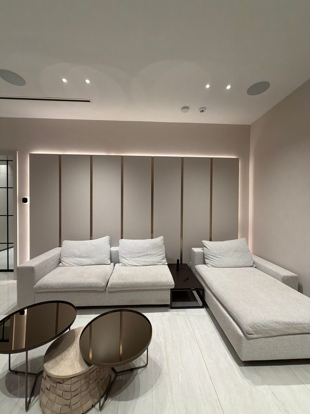 Taupe panel wall with slim vertical metallic reveals and backlit edge glow, light upholstered sofa and chaise, two round dark glass tables on tile