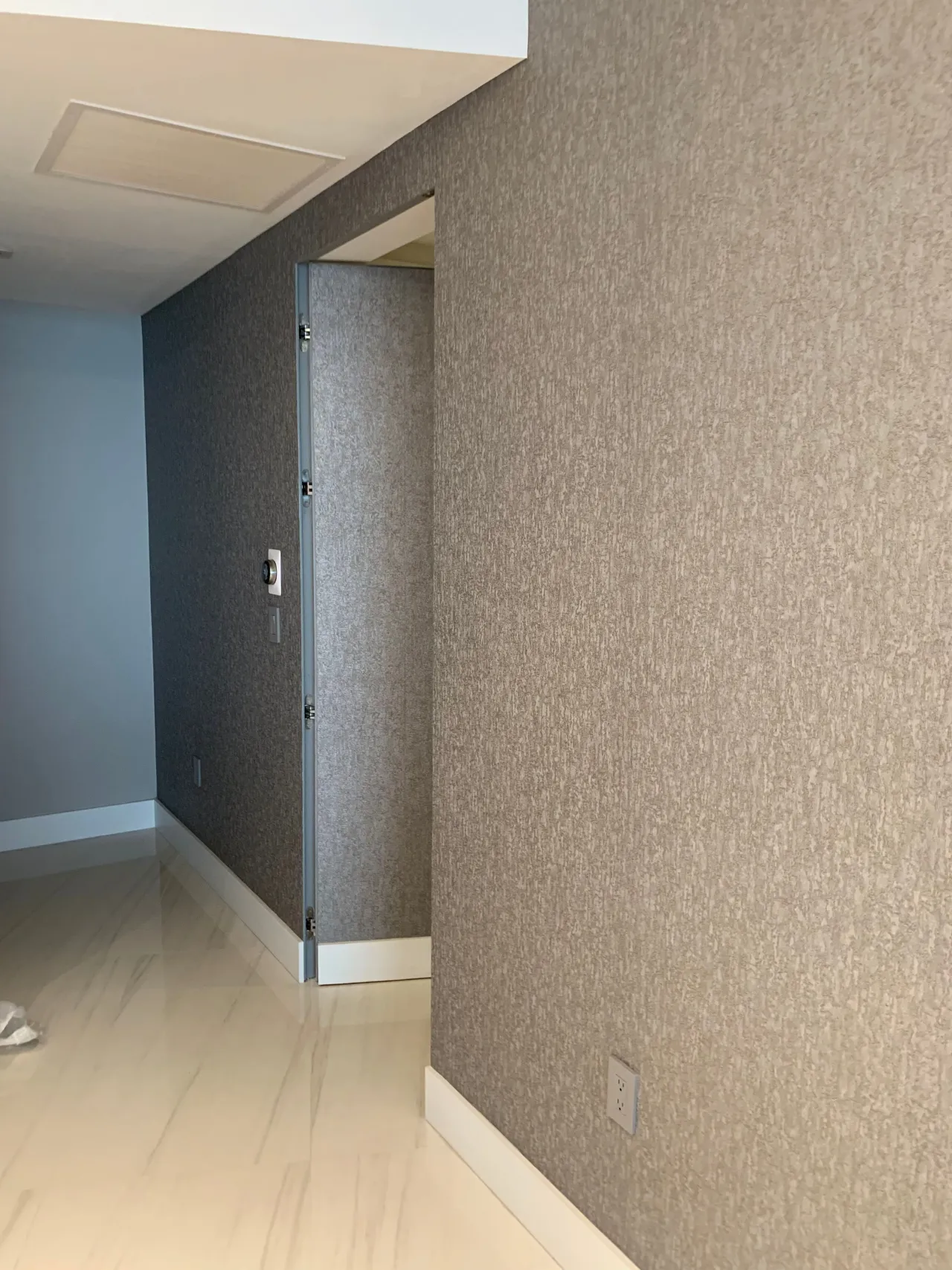 Textured gray fabric-like wall covering with a flush concealed door slightly ajar, revealing a hallway keypad and light interior beyond