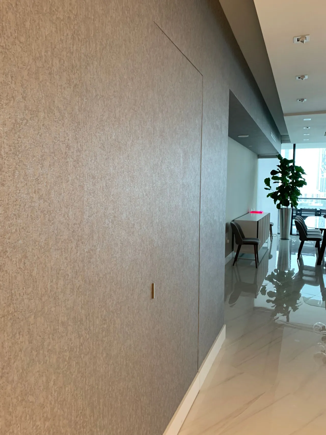 Textured gray wall covering with a flush concealed door outline and small pull, opening onto a bright room with a console table and plant