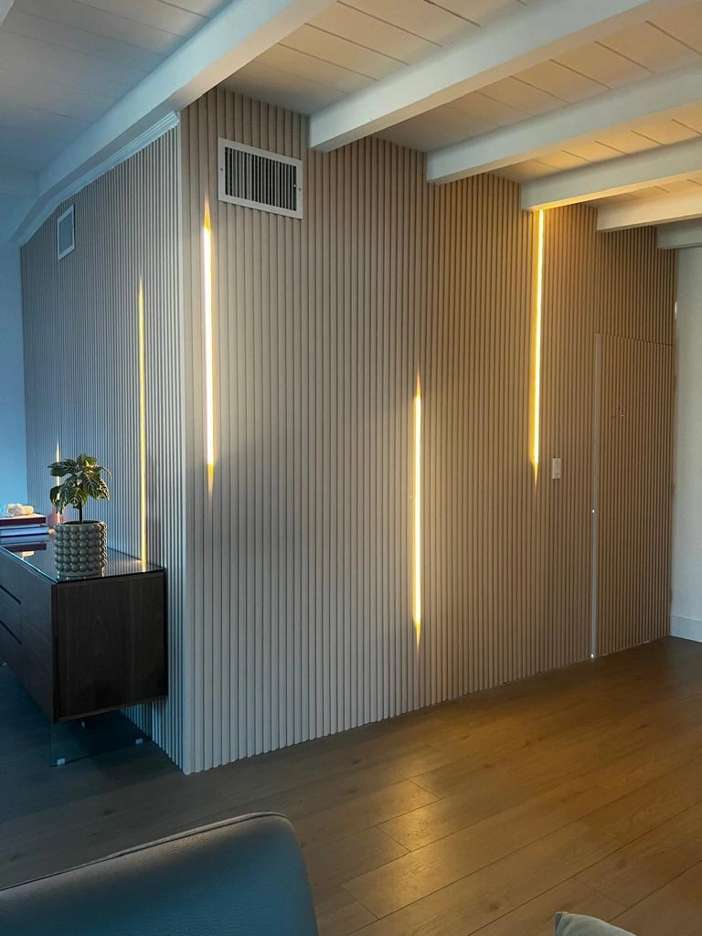 Vertical fluted wall panelling in a greige tone with recessed warm LED strips, a dark floating console at left, medium-tone wood plank floor