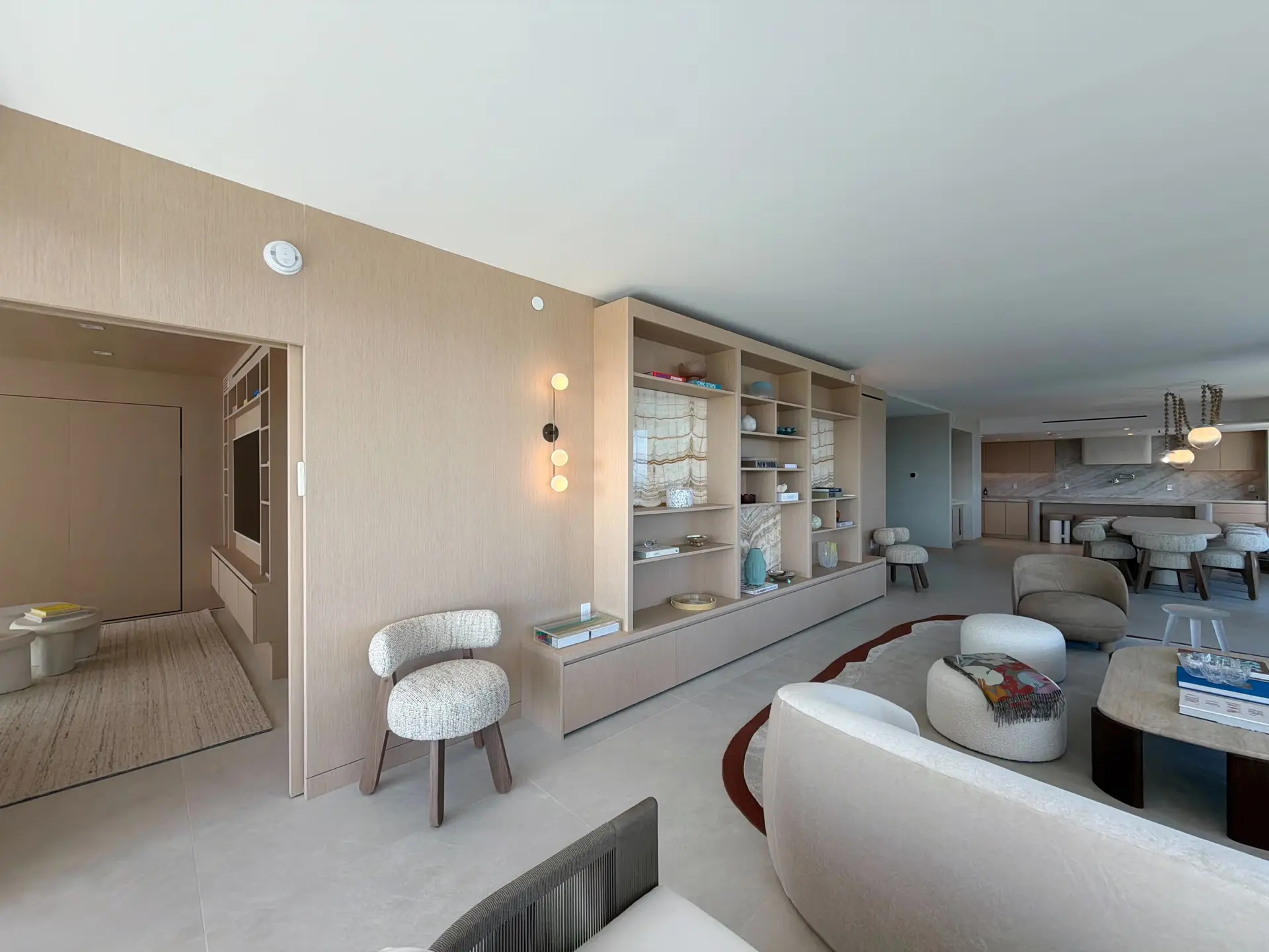 Full-height custom-stained wood veneer built-in shelving wall with backlit stone panels and a media niche, opening to the kitchen and dining area beyond