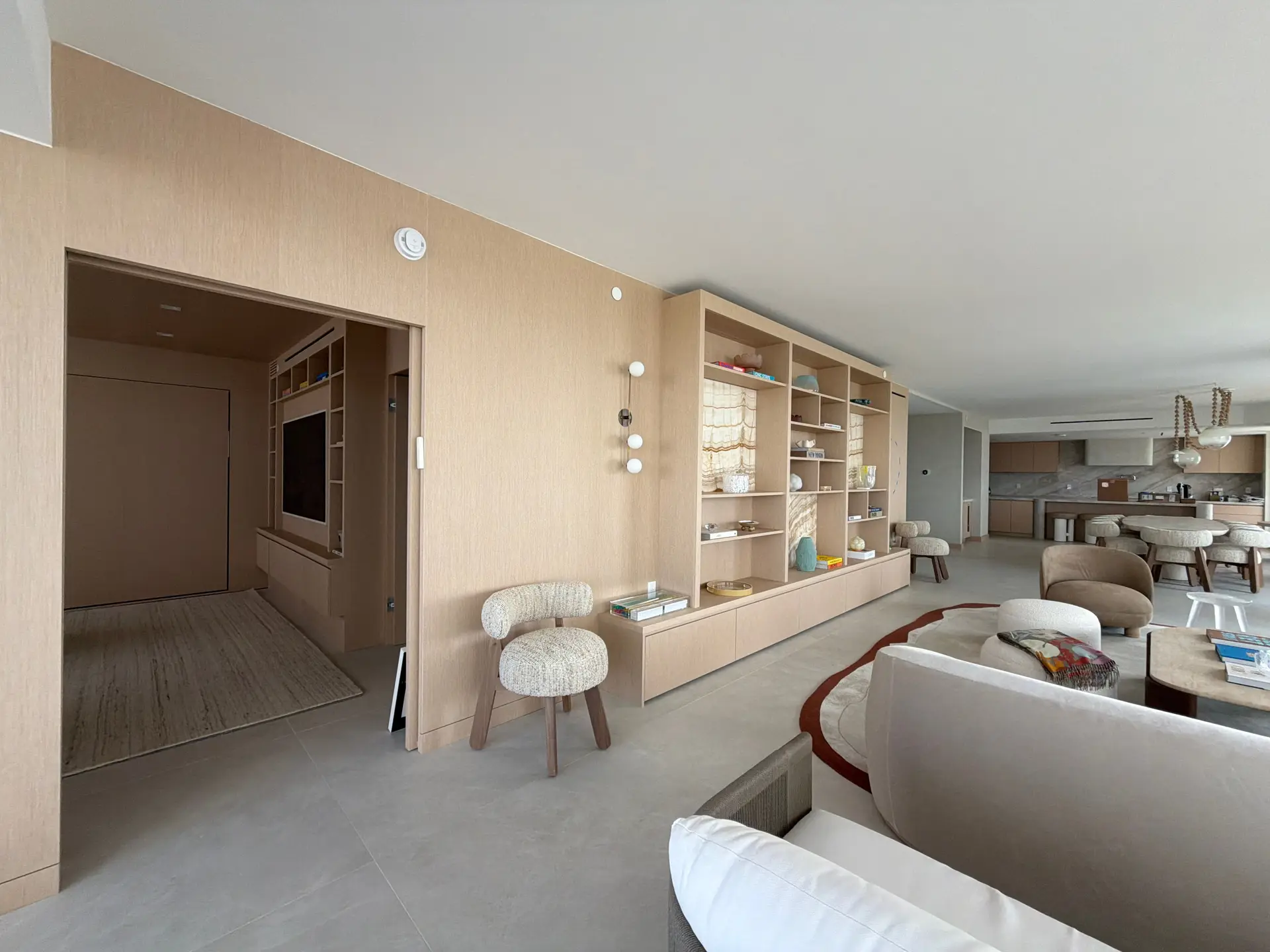 Full-height custom-stained wood veneer media wall with open shelving and a TV niche, opening onto the kitchen and dining area beyond