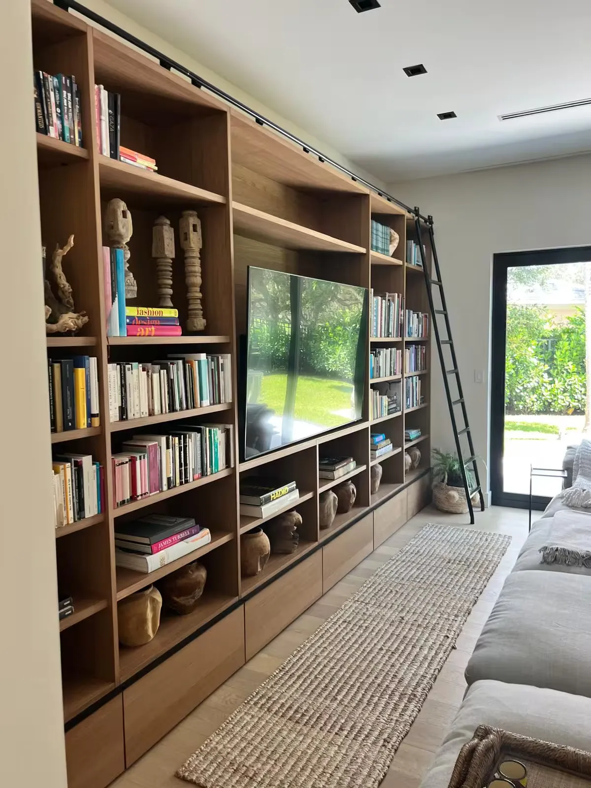 Floor-to-ceiling built-in light wood shelving wall with rolling ladder, TV niche, book displays, and lower drawers